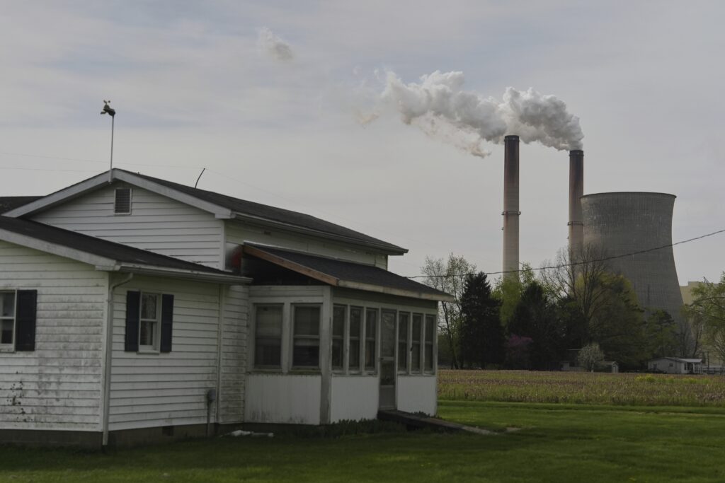 A white house with a small porch and a cow weathervane on the roof, with two industrial smokestacks emitting smoke and a large cooling tower in the background, surrounded by trees and a field of grass.