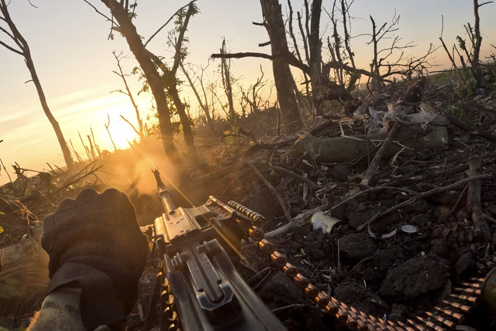 A soldier's gloved hand holding a machine gun with a belt of ammunition in a war-torn landscape, as the sun sets behind bare trees, casting a golden light.