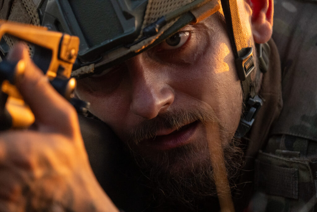 A soldier wearing a tactical helmet is aiming a gun, with intense focus in his eyes. The image is closely cropped, highlighting the soldier's face and the barrel of the gun, capturing a moment of concentration.