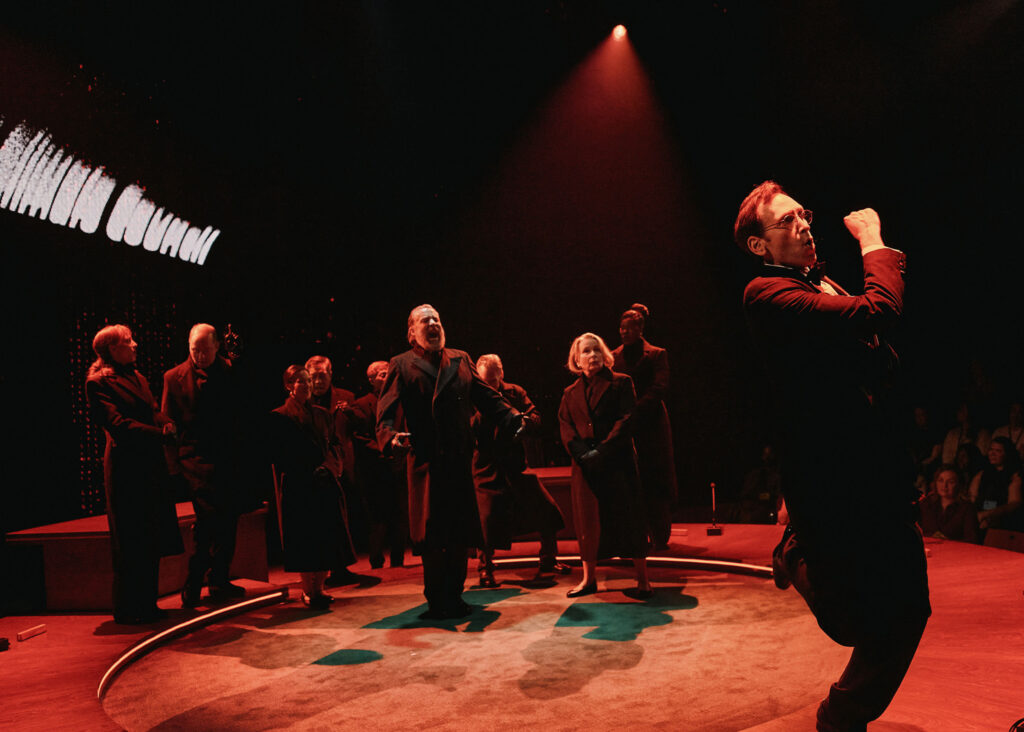 A group of actors on stage under dramatic red lighting, with one performer enthusiastically gesturing in the foreground and others standing and watching in the background.