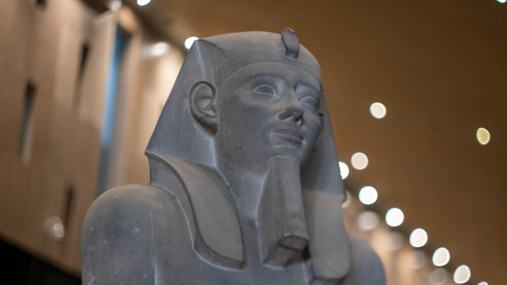 A close-up of an ancient Egyptian statue depicting a pharaoh with a false beard and a headdress, set against a blurred indoor background with soft lighting.