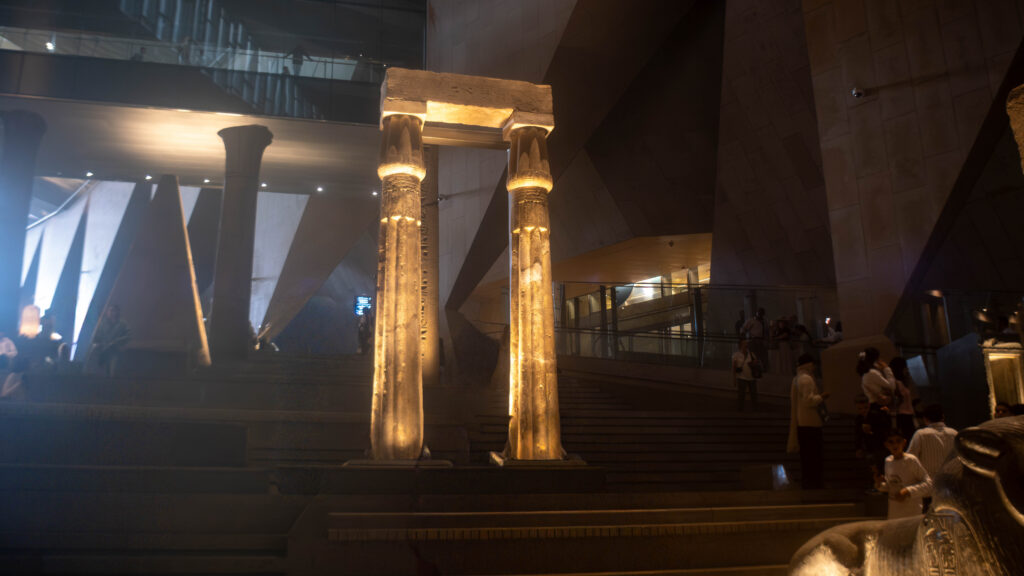 A grand indoor setting with a large, illuminated stone archway supported by two ornate columns, surrounded by people on steps in a modern architectural space.