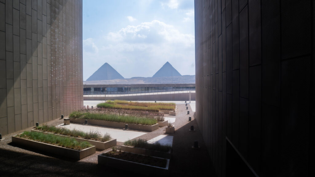 View of two pyramids framed between two modern buildings, with planters filled with greenery in the foreground.