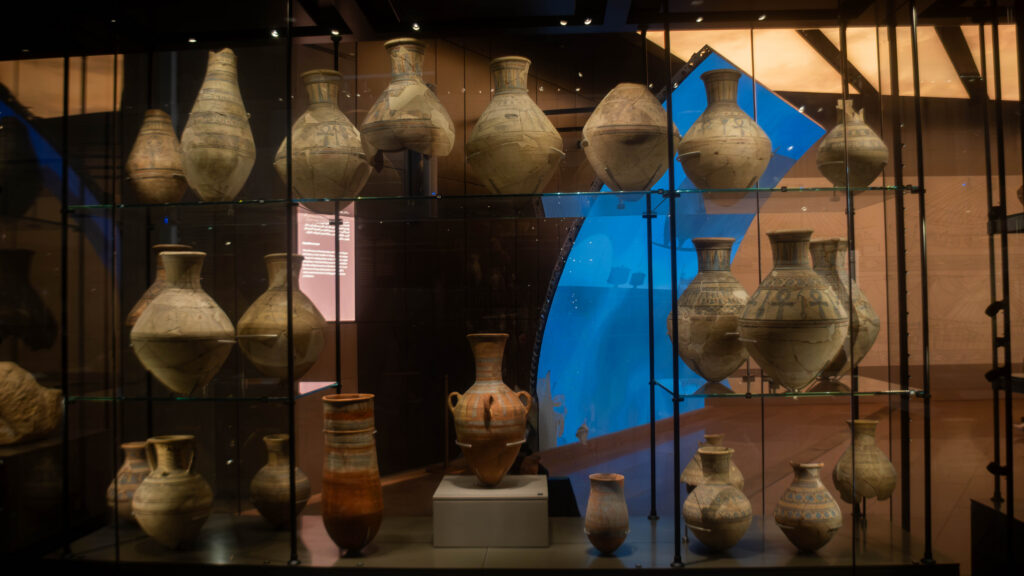 A display case in a museum showcasing a collection of ancient pottery and vases, featuring intricate designs and varying shapes and sizes, with soft lighting highlighting their details.