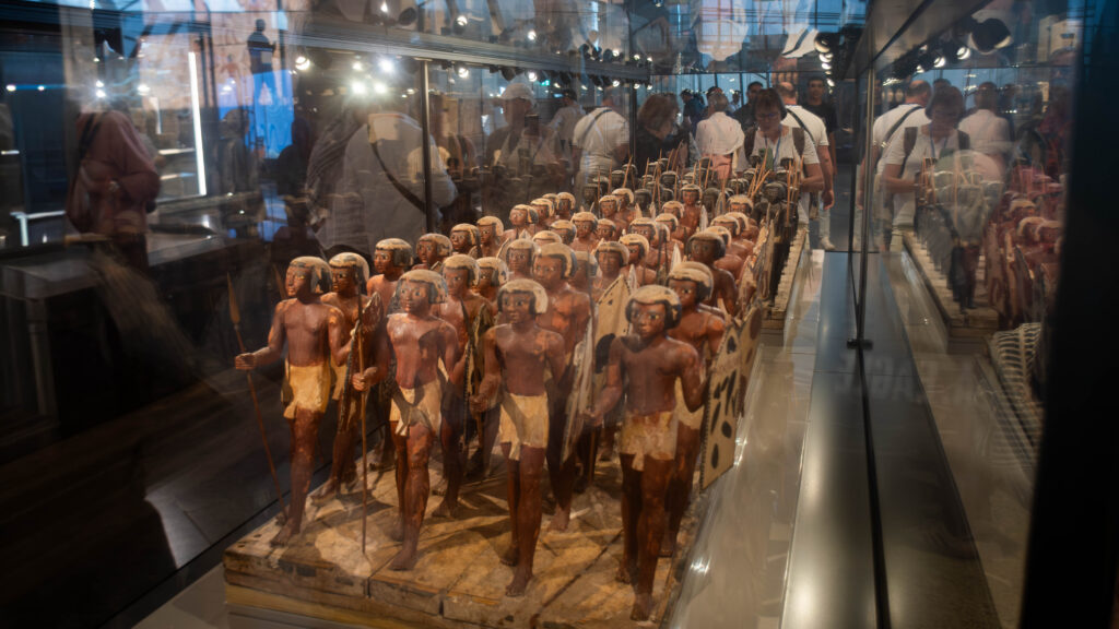 A museum display featuring a group of ancient Egyptian wooden soldier figurines, each holding spears and shields, arranged in rows inside a glass case. Reflections of visitors and overhead lights are visible on the glass surface.