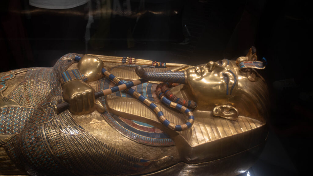 A golden sarcophagus with intricate detailing and blue ceramic inlays, depicting an ancient Egyptian figure with crossed arms and a ceremonial beard.