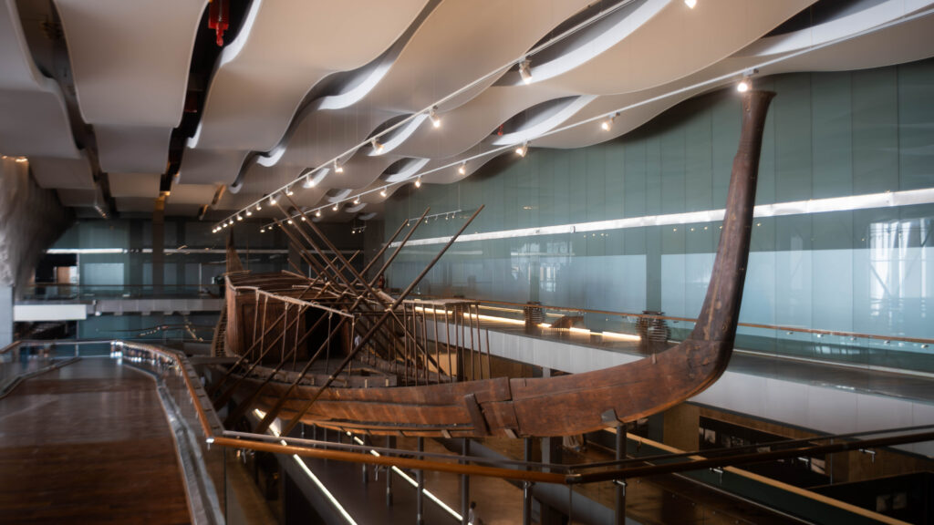 A wooden ship display in a modern museum interior, featuring curved ceilings and glass walls.