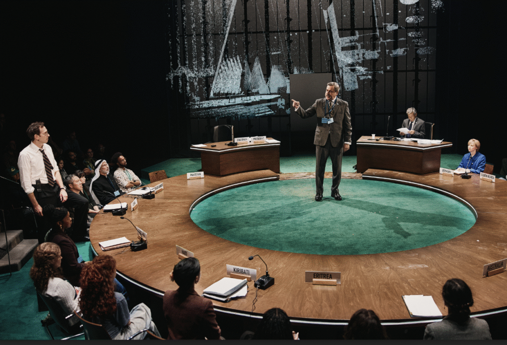 A group of people seated around a large circular conference table, with nameplates for various countries. A man stands in the center, speaking, while others listen attentively. The setting resembles a formal international meeting, possibly simulating a United Nations or diplomatic assembly.