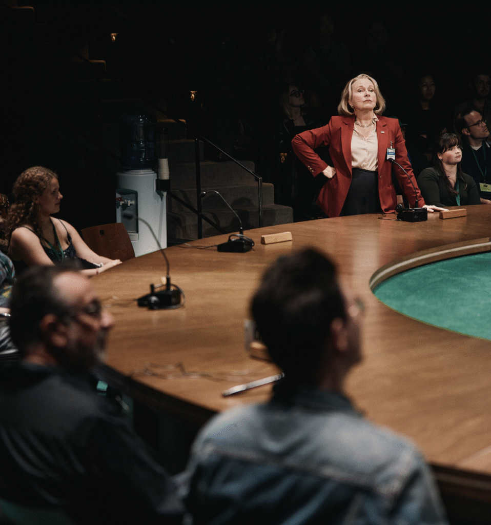 A woman in a red blazer stands assertively at a round conference table, surrounded by people seated and watching her, in a dimly lit room.