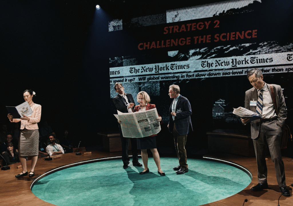 A group of actors perform on stage with a backdrop displaying headlines from The New York Times and the phrase