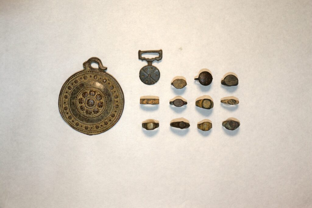 An assortment of ancient metallic artifacts including a large circular medallion with intricate designs, a smaller circular pendant with a geometric pattern, and a collection of decorative rings arrayed in rows on a plain background.