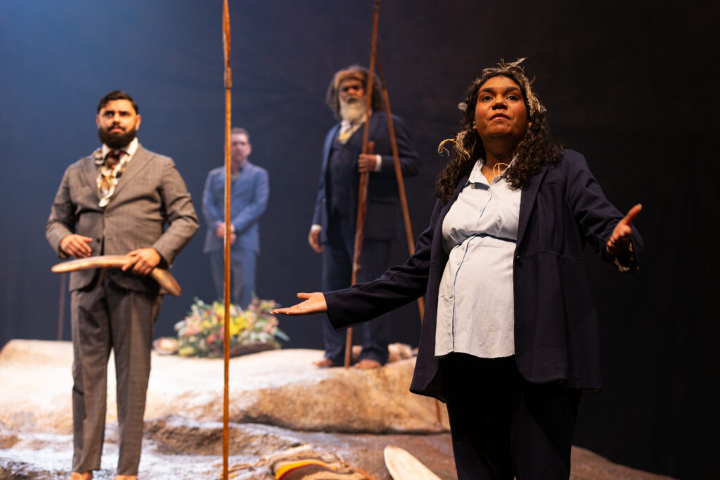 A woman in a suit speaks passionately on stage, holding out her arms, with three men standing behind her, one holding a stick, in a dramatic setting with rocks and traditional objects.