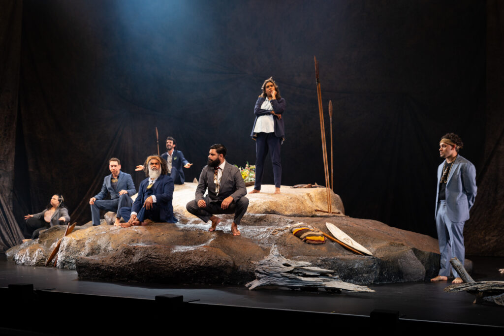 A group of people in formal attire perform on stage, standing and sitting on a large, rocky set, with spears and driftwood nearby.