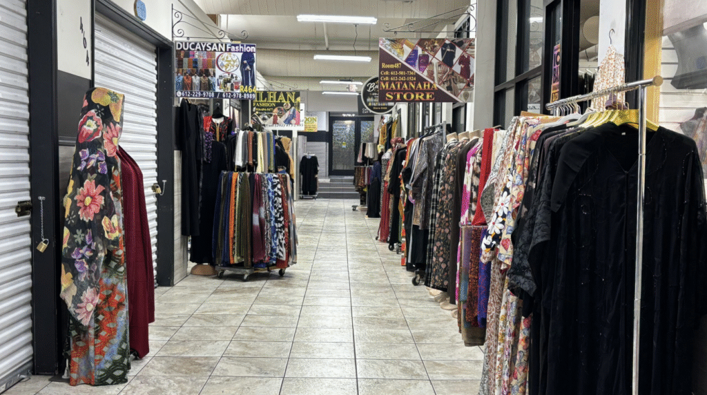 An indoor clothing marketplace with racks of colorful clothes lining both sides of a tiled walkway. Several signs for different fashion stores hang from the ceiling.