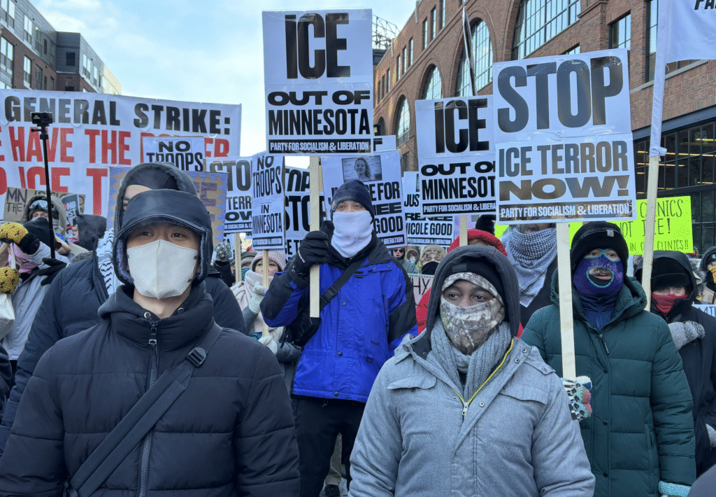 A group of people are gathered at a protest, holding signs with messages such as 'ICE OUT OF MINNESOTA', 'STOP ICE TERROR NOW!', and 'GENERAL STRIKE'. Many participants are bundled in winter clothing and face masks, standing in front of a brick building.
