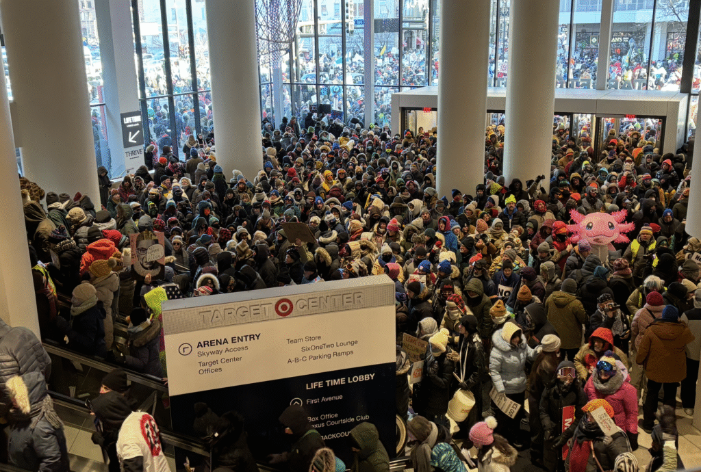 A large crowd of people bundled up in winter clothing fills the interior of the Target Center entrance. Many are holding signs, and there's a prominent inflatable pink creature among the crowd. Outside through the glass windows, more people are visible, indicating a large gathering or event.