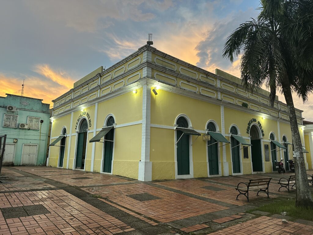 A yellow historic building labeled 'Mercado Velho' with arched green doors and windows, surrounded by a paved area with benches and a palm tree, under a cloudy sunset sky.