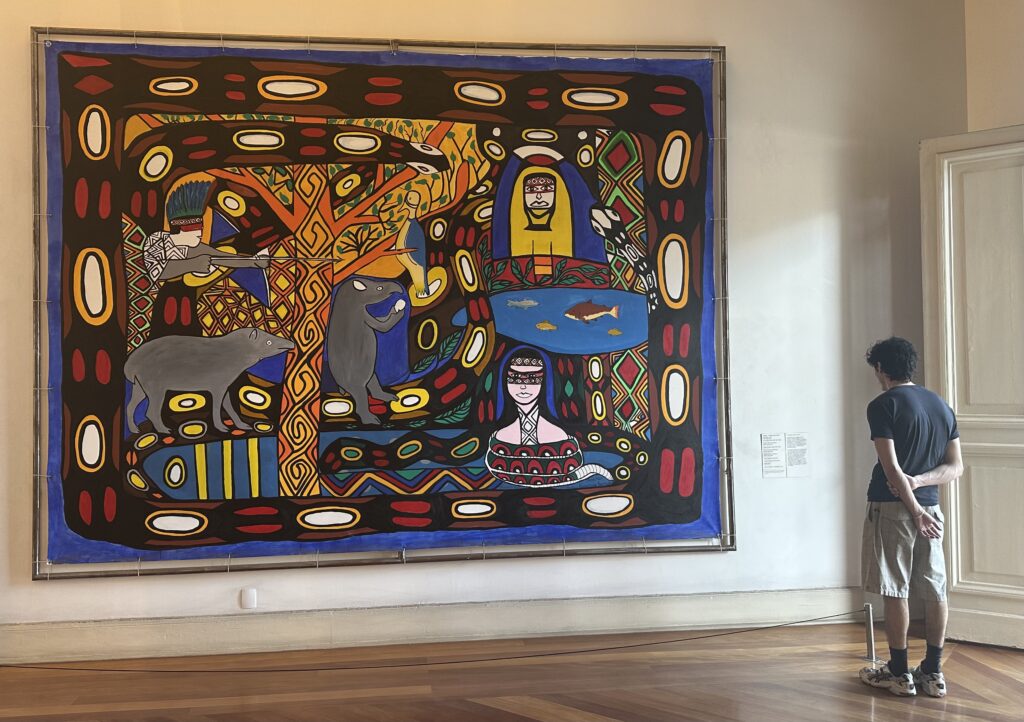 A person stands in an art gallery observing a large, colorful painting that features vivid patterns, animals, and human figures in an abstract style. The painting includes elements like a tree, animals, and geometric designs, framed by bold colors.