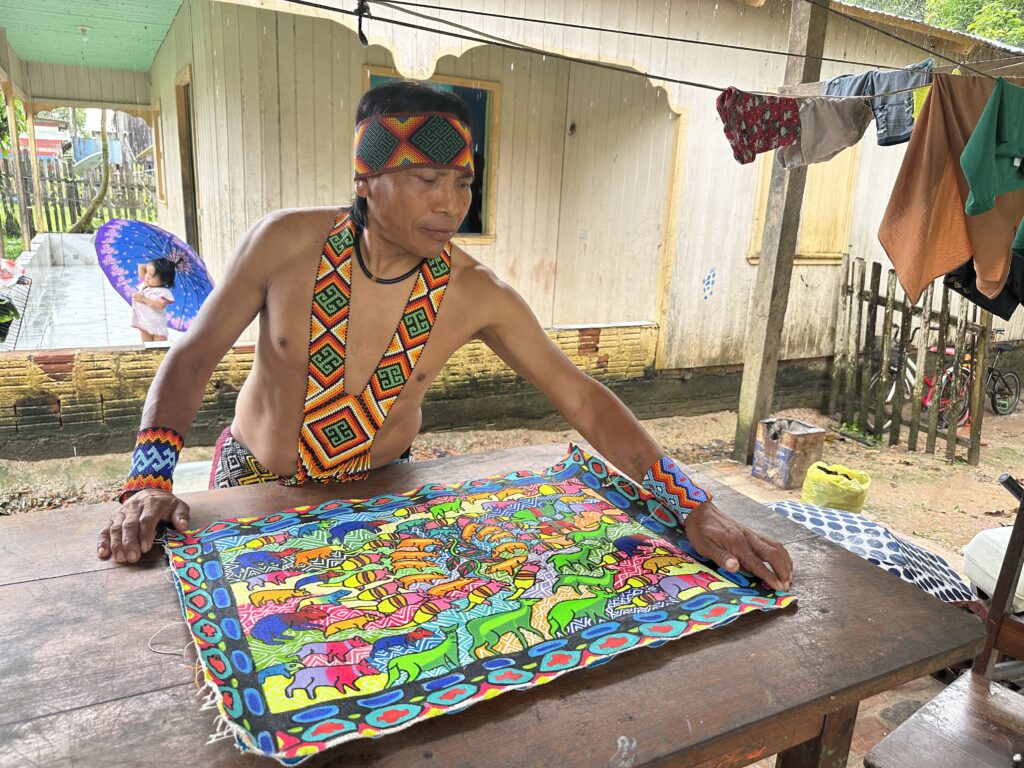 A man wearing traditional attire with vibrant beadwork arranging a colorful patterned cloth on a wooden table, with clothes hanging on a line and a child holding an umbrella in the background.
