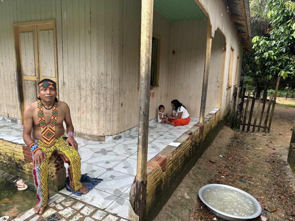 A man in traditional attire with colorful patterns sits outside a rustic wooden house. In the background, a woman, and a child are sitting on the porch. The scene includes a dirt path and a metal basin filled with water in the foreground.