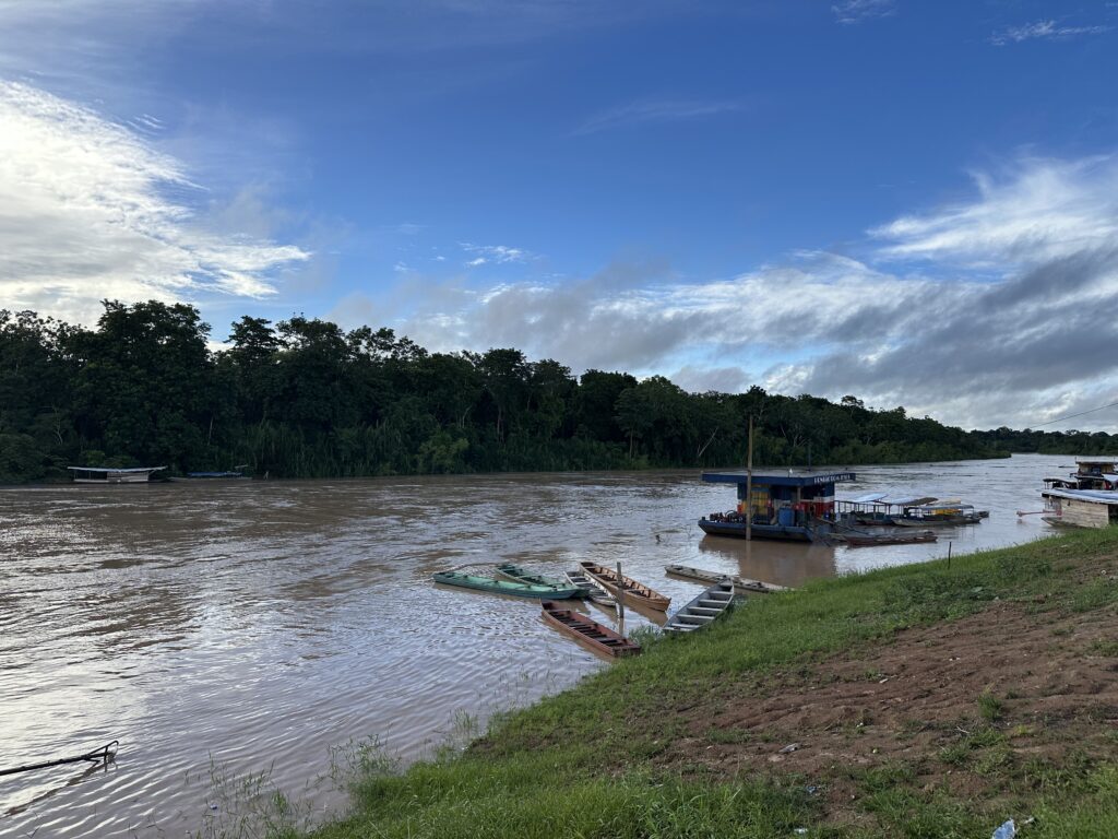 A river with several boats docked near the shore, surrounded by lush green trees on the opposite bank, under a partly cloudy sky.
