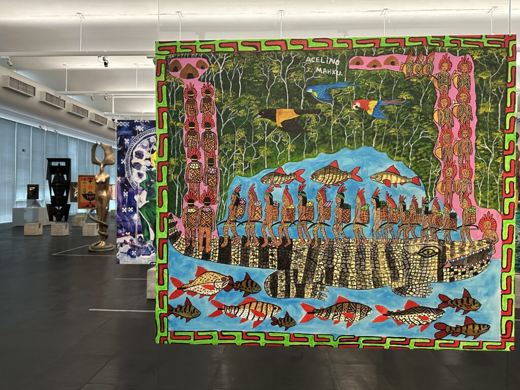 A colorful painting displayed in a gallery, depicting people standing on a large striped crocodile surrounded by fish, with a forest background. Other artworks are visible in the gallery space.