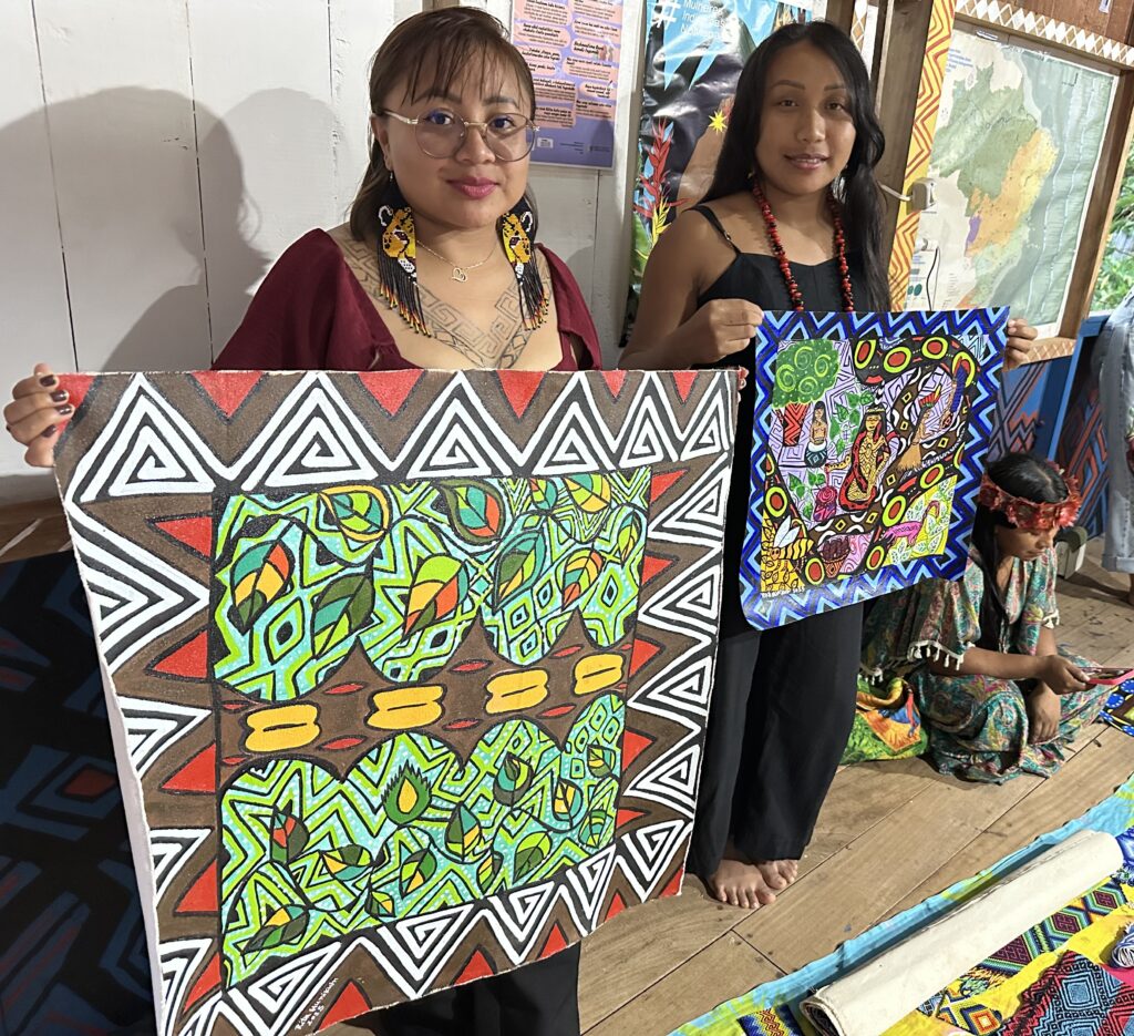 Two women proudly display colorful, geometric paintings with indigenous motifs, standing inside a room with another person seated on the floor in traditional attire.