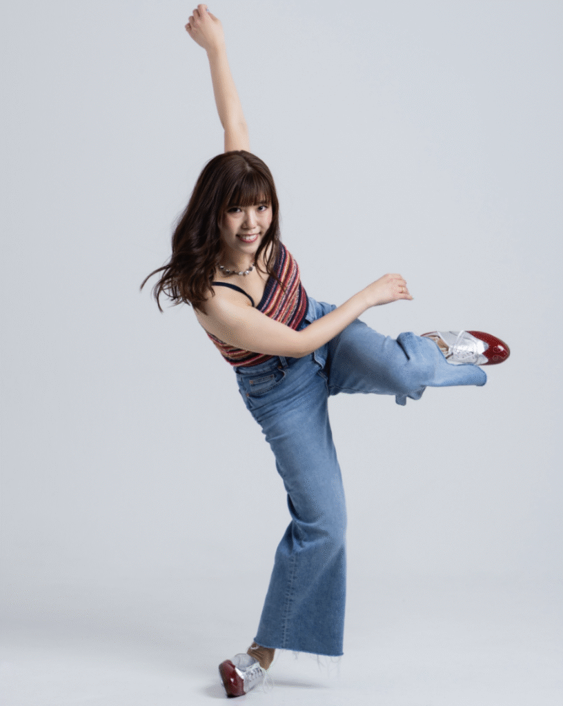 A person wearing a colorful striped tank top and blue jeans, performing a dynamic dance move with one arm raised and one leg lifted.