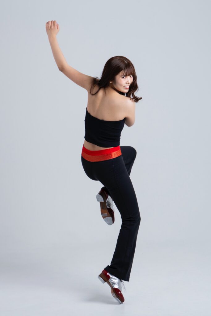 A person wearing a black sleeveless top and black pants with a red waistband, jumping while dressed in tap shoes, set against a plain background.