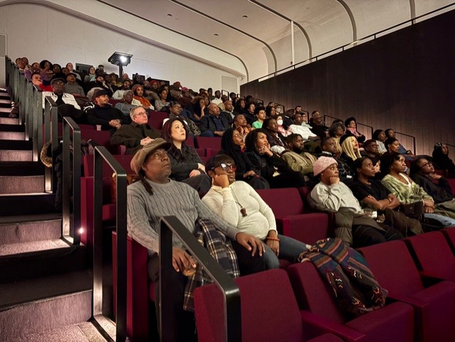 A diverse group of people seated in a large, modern auditorium, attentively watching a presentation or performance. The auditorium is filled with rows of burgundy-colored seats.