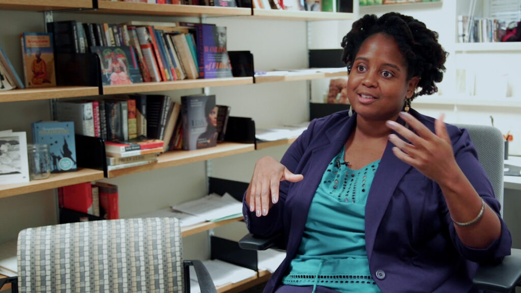 A person speaking animatedly in an office with bookshelves filled with books in the background.