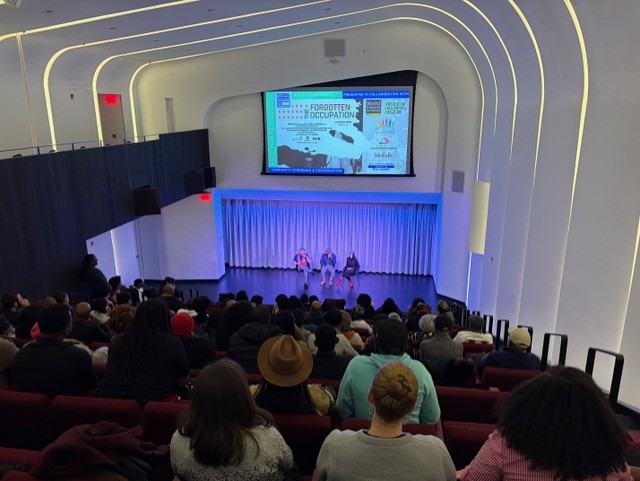 An auditorium filled with people watching a panel discussion. The stage hosts three speakers seated under a large screen displaying the title 'Forgotten Occupation.' The room has modern architectural design with curved lines and overhead lighting.