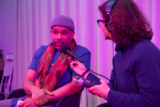 A man wearing a gray knit hat and a colorful scarf is speaking into a microphone held by another person, who is wearing headphones and holding recording equipment. The setting has a purple lighting ambiance.