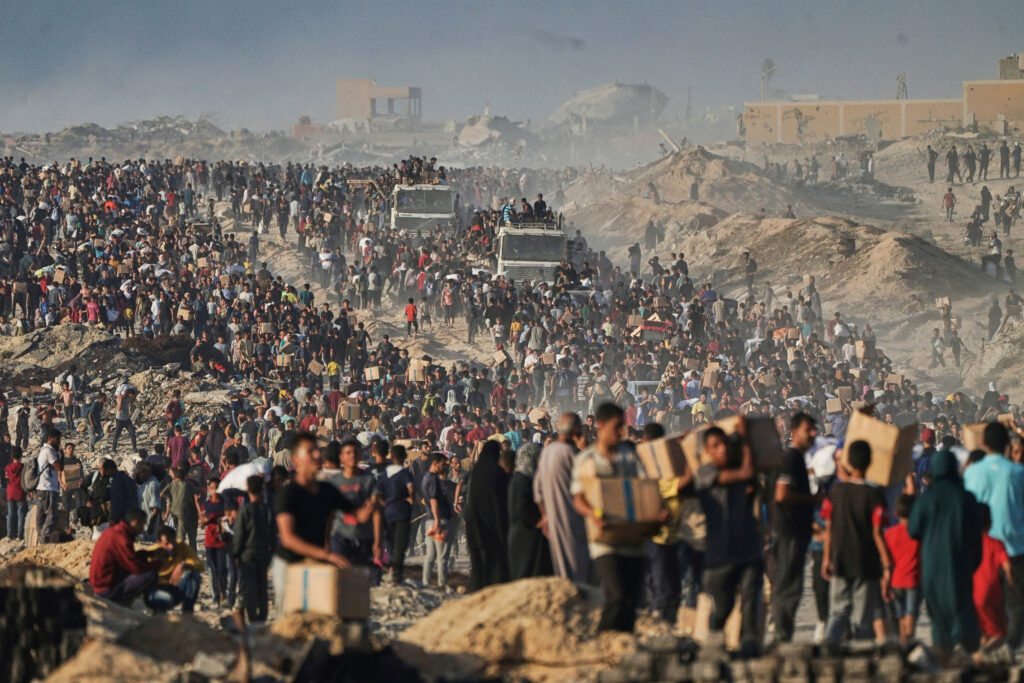 A large crowd of people carrying belongings walks through a dusty landscape, with buildings and vehicles visible in the distance.