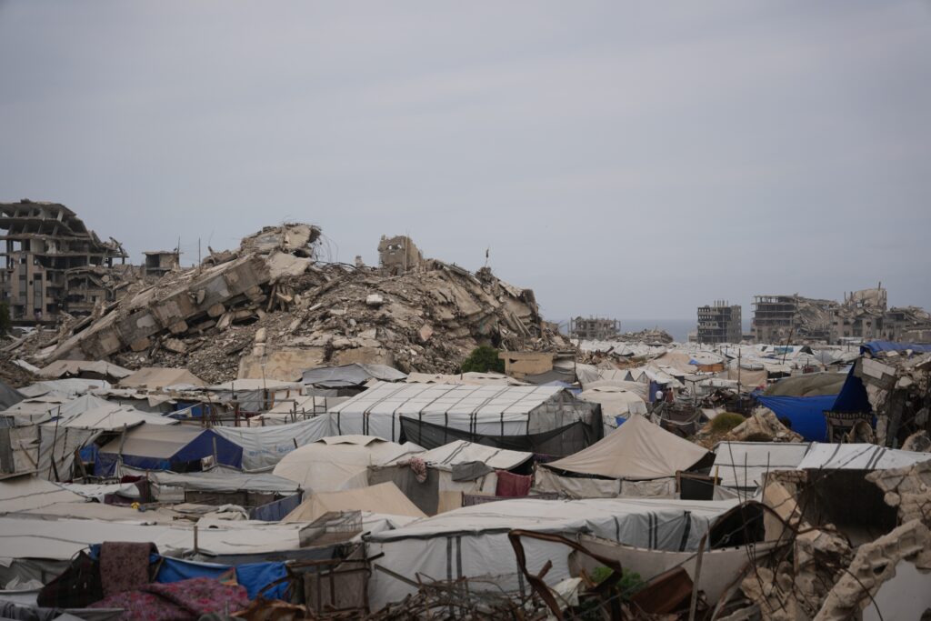 A large area filled with makeshift tents is set against a backdrop of heavily damaged, collapsed buildings.