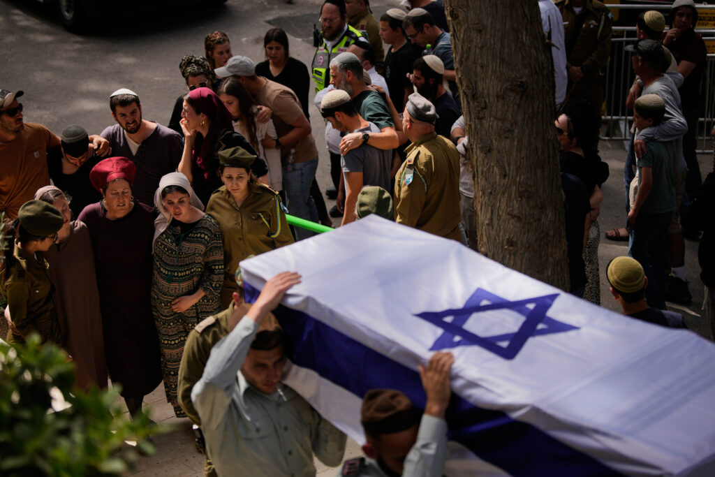 A group of mourners, including soldiers, attend a funeral procession with a flag-draped coffin bearing the Star of David.