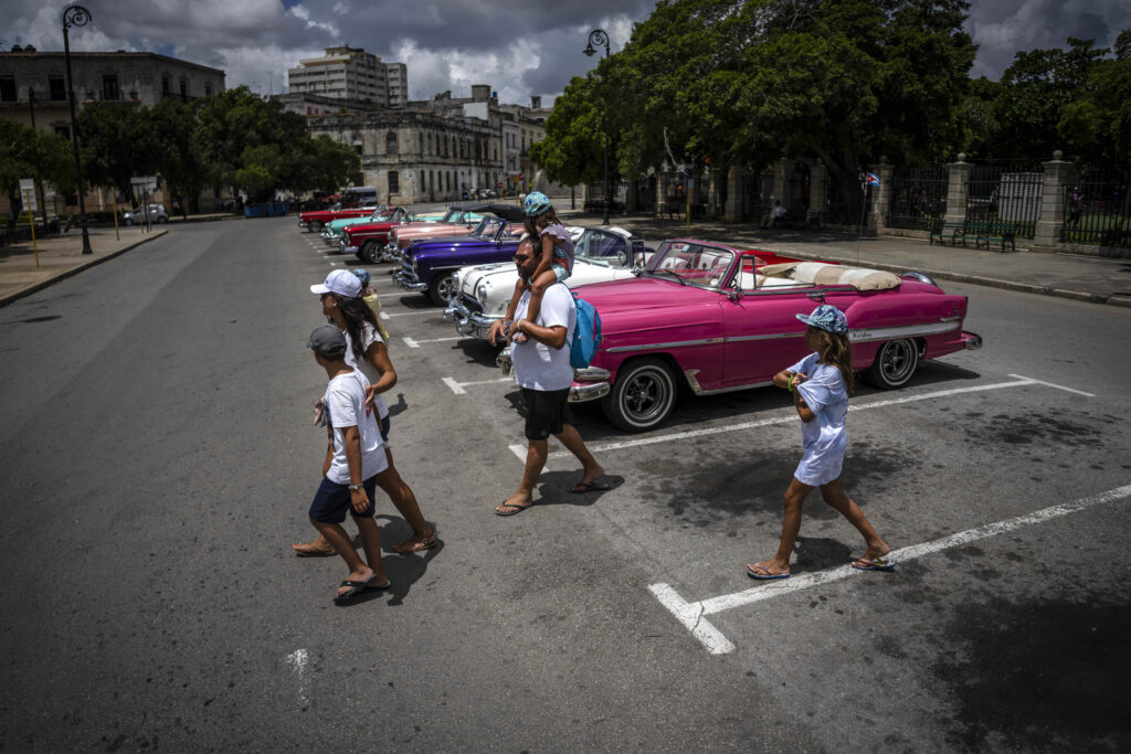 A family crosses a street in front of a lineup of colorful classic cars parked in an urban setting under a partly cloudy sky.