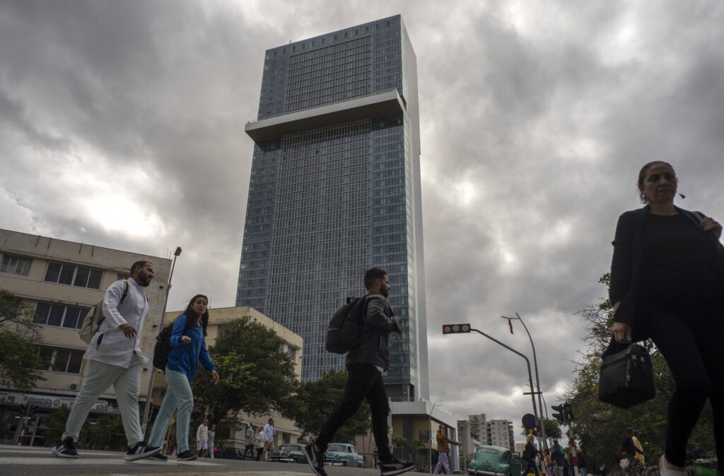 People walking near a tall, modern skyscraper under a cloudy sky.