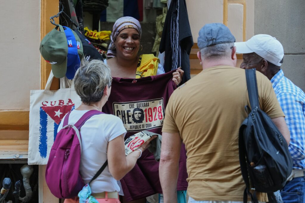 A street vendor shows a Che Guevara shirt to three tourists near bags with a Cuban flag design.