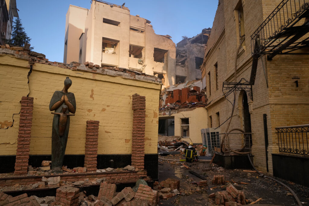 A damaged building surrounded by rubble, with a standing statue and a person in protective gear in the background.