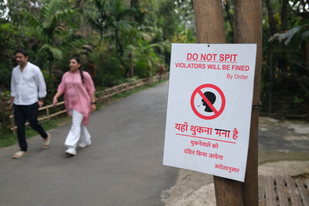 Sign warning against spitting with a fine, featuring text in English and another language, near a path in a park.