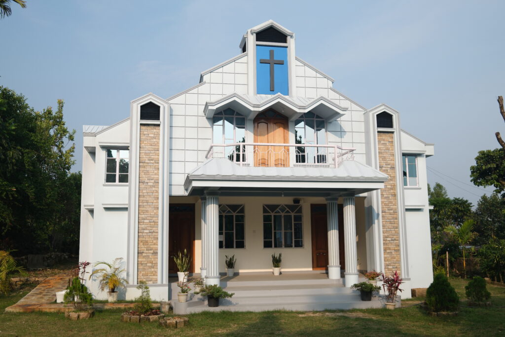 A white church with a cross above its entrance, featuring columns and a small garden in front.