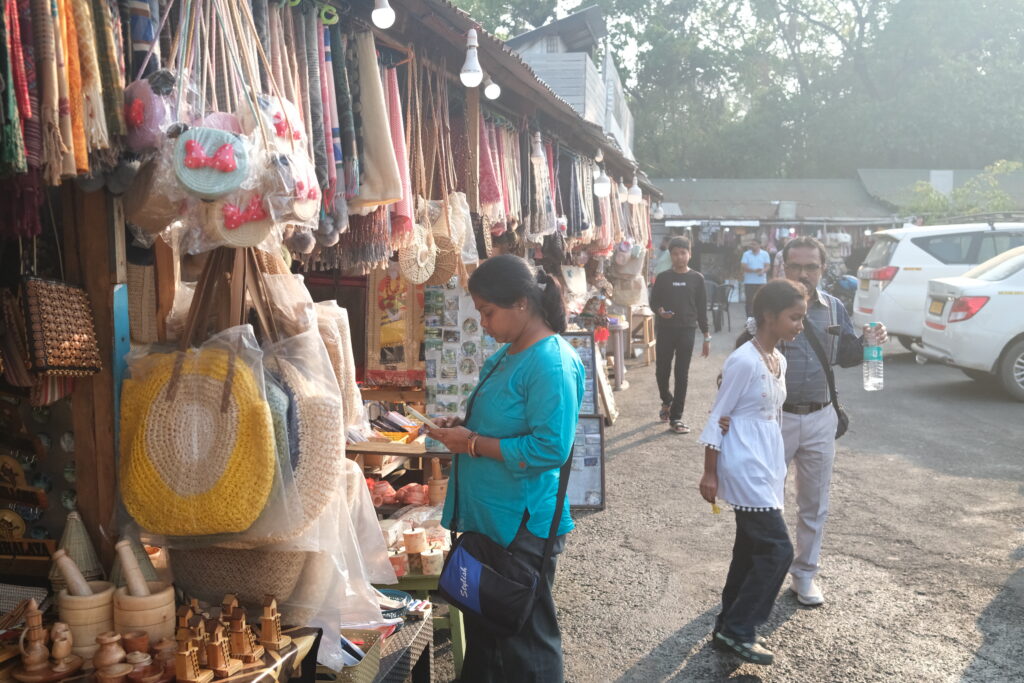 A woman shops at a street market stall displaying bags and craft goods, with other people browsing nearby.