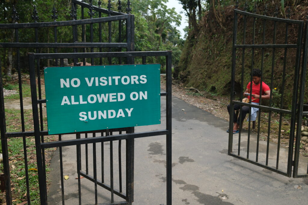 A sign reads "No visitors allowed on Sunday" near a man in a red shirt sitting by a gate, surrounded by greenery.
