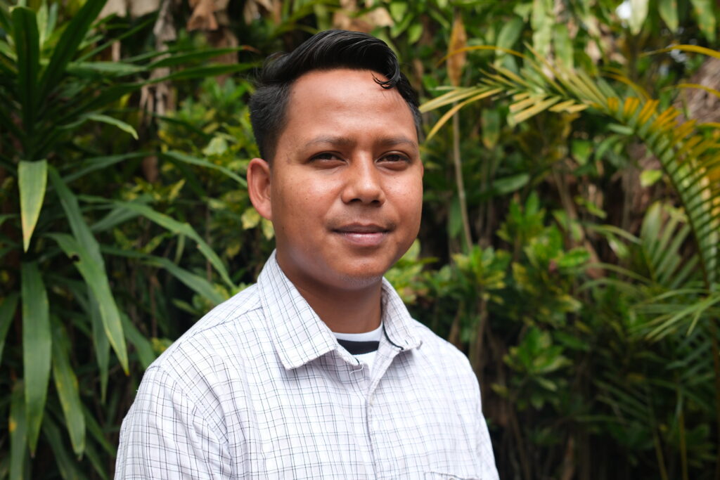 A person wearing a checked shirt is standing in front of lush green foliage.