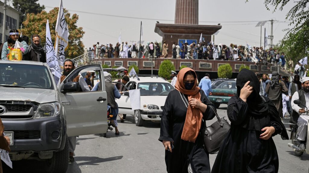 Two women in headscarves walk through a crowded street in Kabul as Taliban fighters with white flags ride in trucks nearby