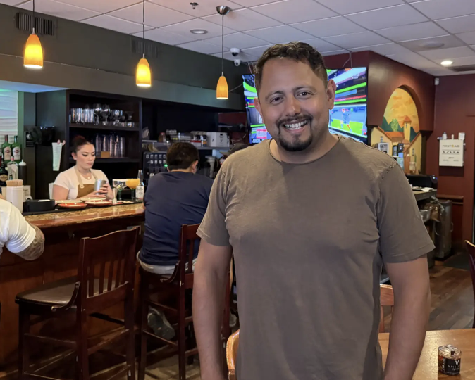 A smiling man stands in a casual restaurant or bar, with a bartender and patrons visible in the background