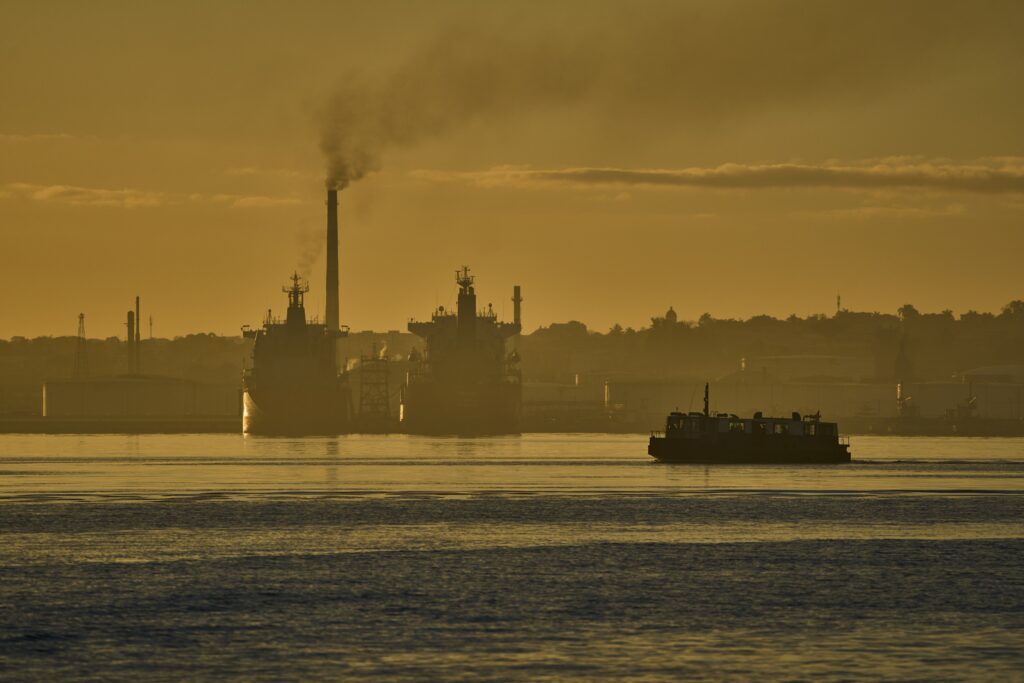 Industrial waterfront at dusk with a smoking smokestack, silhouetted ships, and a barge crossing a hazy golden harbor
