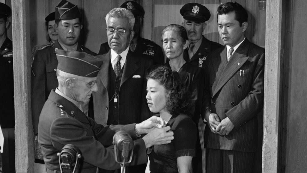 A U.S. Army officer pins a medal on a Japanese American woman at a ceremony, with family members and military personnel looking on in the background