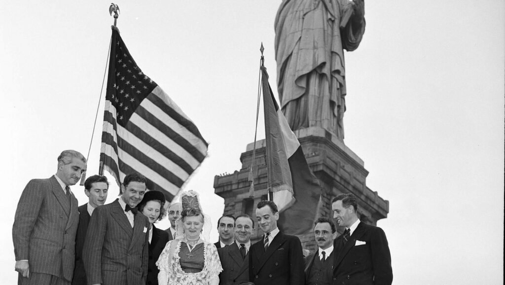 A group of people pose before the Statue of Liberty with American and French flags, one woman wearing traditional folk dress and lace headpiece